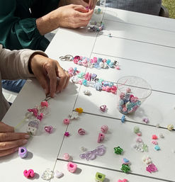 Two people crafting pastel bead keychains on a sunlit white slatted outdoor table, with scattered heart, star and alphabet charms