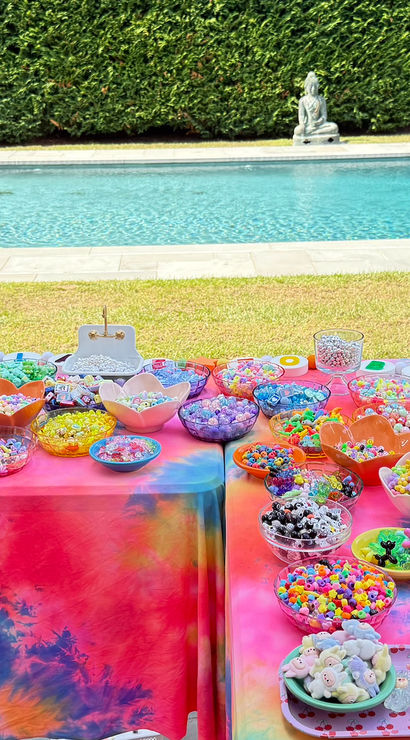 Poolside backyard craft spread with rainbow tie-dye tablecloths covered in bowls of colorful beads, charms, strings and jewelry-making tools beside a blue swimming pool and manicured hedge.