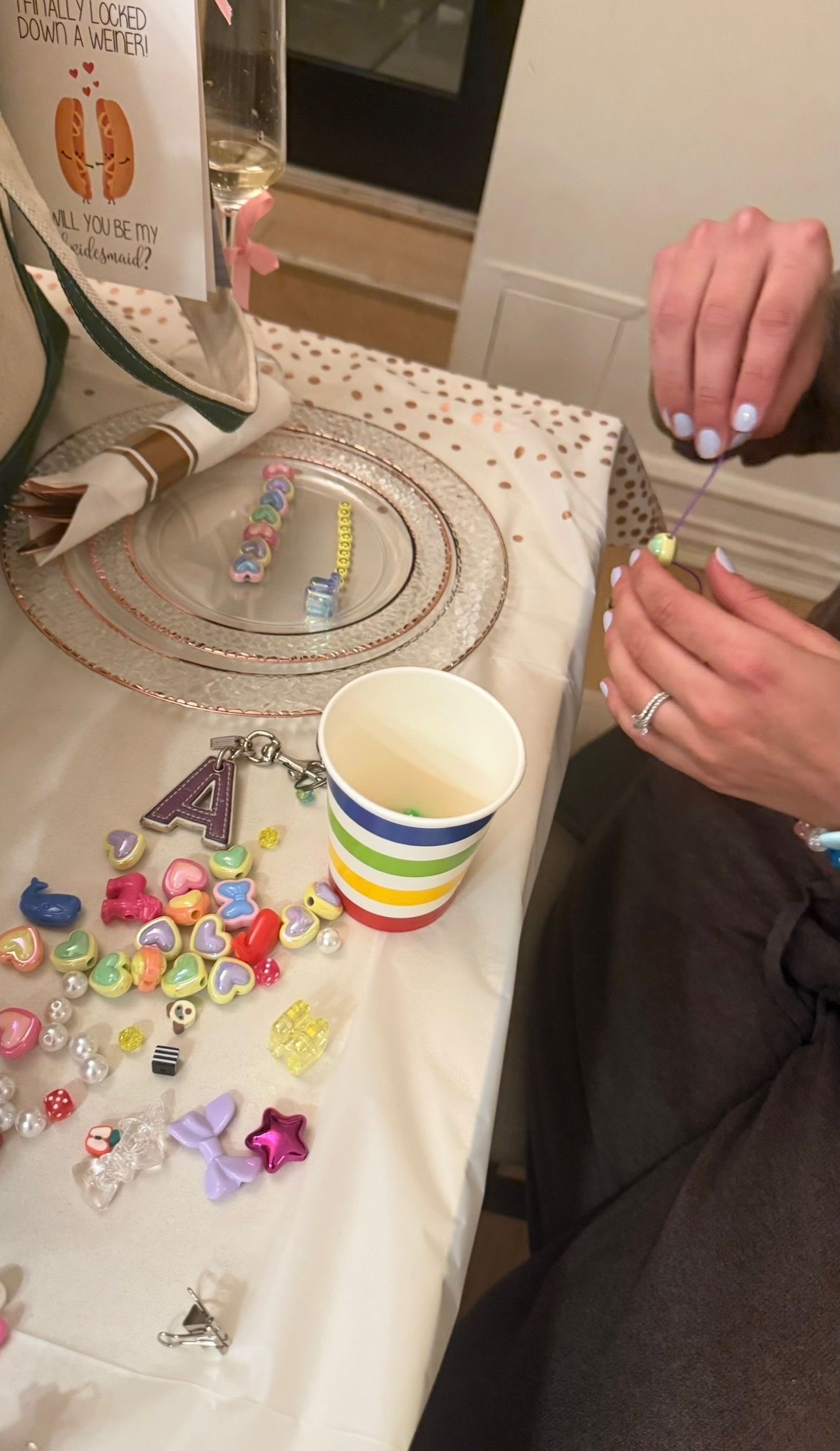 Hands stringing pastel heart beads at a bridal shower craft table with a rainbow-striped cup, scattered colorful charms and rose-gold charger plate.