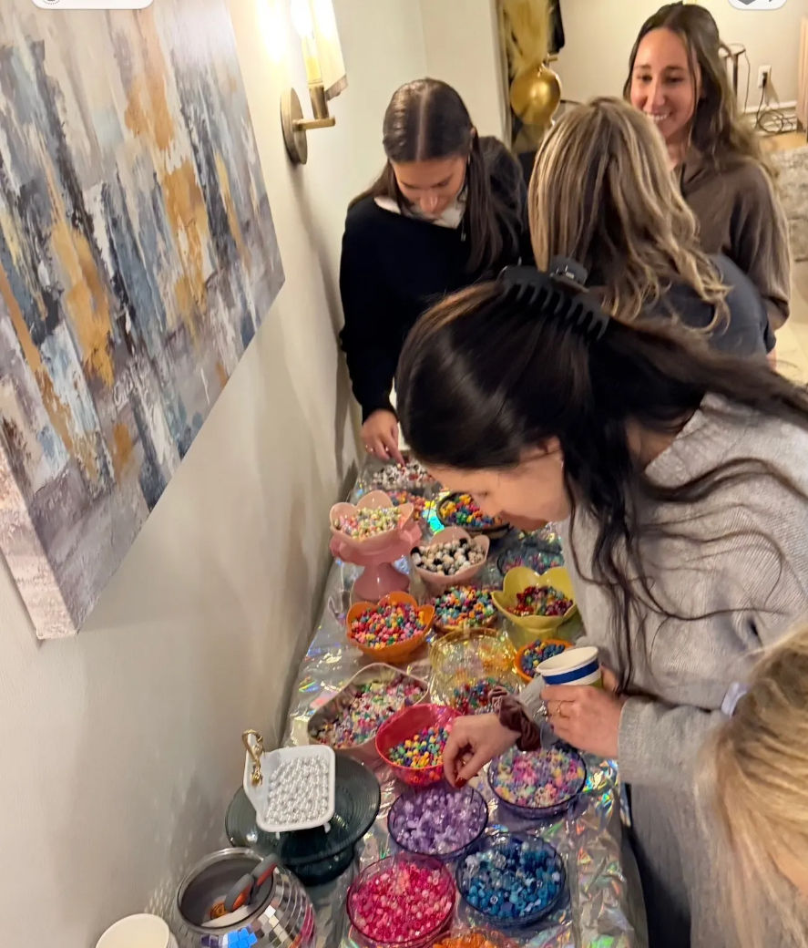 Guests at an indoor party selecting colorful beads and candies from a long buffet of bowls — a DIY bead-craft and candy bar setup on a living-room table.