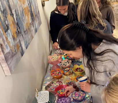Guests at an indoor party selecting colorful beads and candies from a long buffet of bowls — a DIY bead-craft and candy bar setup on a living-room table.