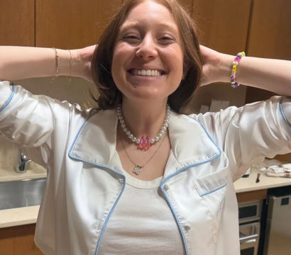 Beaming woman in a white pajama-style top standing in a home kitchen with wood cabinets, hands behind her head, wearing a pearl necklace with a pink charm and a colorful beaded bracelet.