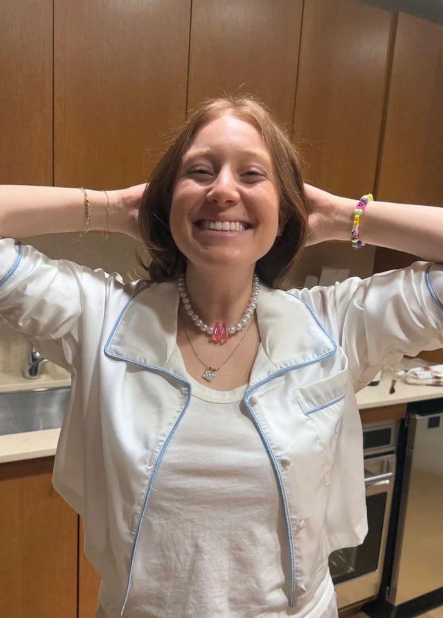 Beaming woman in a white pajama-style top standing in a home kitchen with wood cabinets, hands behind her head, wearing a pearl necklace with a pink charm and a colorful beaded bracelet.