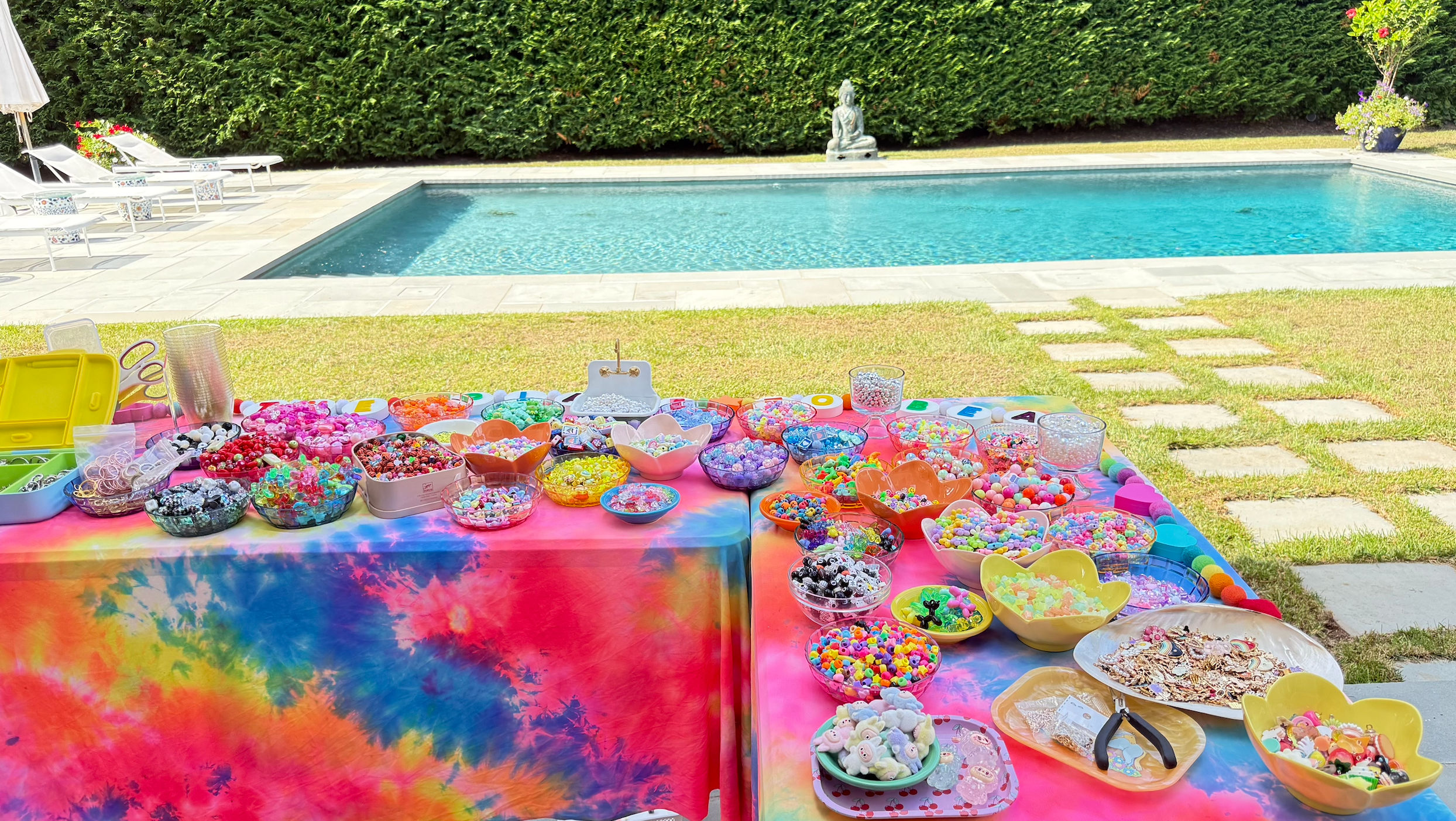 Poolside backyard craft spread with rainbow tie-dye tablecloths covered in bowls of colorful beads, charms, strings and jewelry-making tools beside a blue swimming pool and manicured hedge.