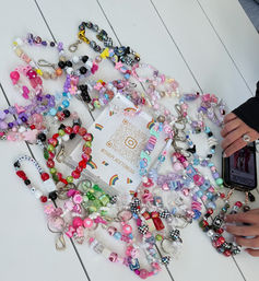 Colorful handmade beaded bracelets and keychains with charms scattered across a white wooden table around a QR promo card, hands holding a phone at the edge
