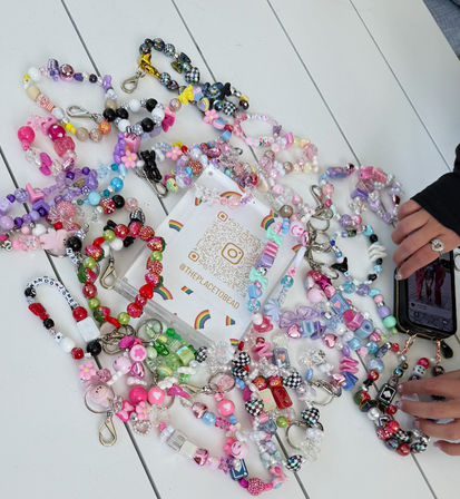 Colorful handmade beaded bracelets and keychains with charms scattered across a white wooden table around a QR promo card, hands holding a phone at the edge