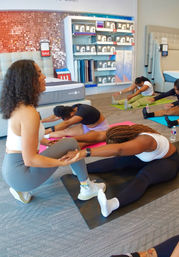 Group yoga/stretching class in a mattress showroom — participants on mats doing seated forward stretches near bedding displays and sale signage