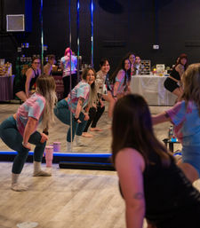 Group of women in a lively dance fitness class at an indoor mirrored studio, squatting and smiling near chrome poles while wearing colorful tie-dye tops and leggings on a wood floor