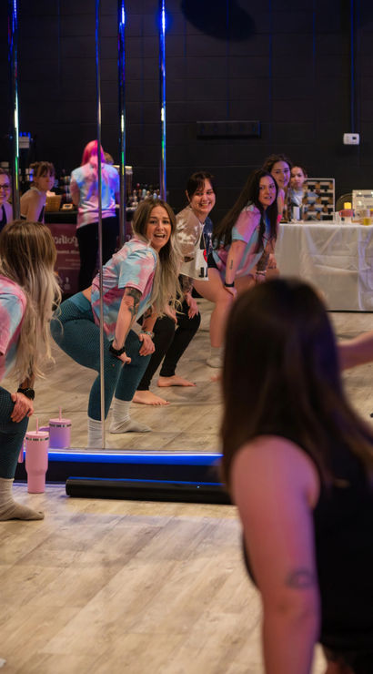 Group of women in a lively dance fitness class at an indoor mirrored studio, squatting and smiling near chrome poles while wearing colorful tie-dye tops and leggings on a wood floor