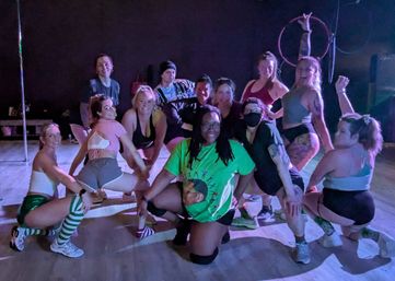 Energetic group of fitness class participants posing in a dimly lit dance studio with a pole and aerial hoops, wearing colorful athletic shorts, crop tops, knee pads and striped socks, smiling and crouching in a playful group pose.