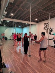 Group pole-fitness class in an urban dance studio — diverse adults practicing around chrome poles on polished wood floors under an exposed-beam ceiling with a wall mural.