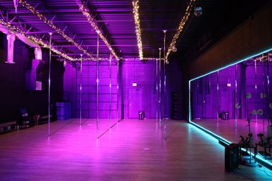Empty pole fitness studio interior with polished chrome poles, floor-to-ceiling mirror, purple neon and fairy lights, warm wood floor.