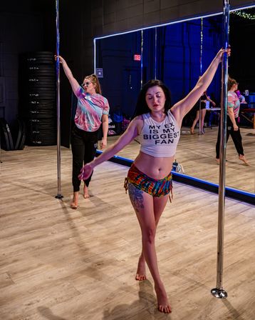 Two women in a pole fitness studio practicing by mirrored walls and wood floors — foreground dancer in a 'MY EX IS MY BIGGEST FAN' crop top and colorful shorts holds a chrome pole, eyes closed.