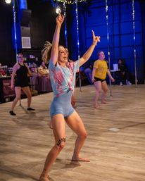 Energetic pole dance fitness class in an indoor studio — woman in tie-dye top and blue shorts leads an upbeat routine with arms raised on a wooden floor under blue lights.