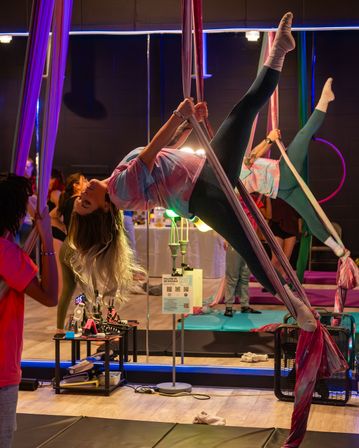 Person defying gravity in an upside-down aerial silks pose inside a vibrant indoor aerial yoga/fitness studio with mirrored wall, classmates, mats and colorful equipment
