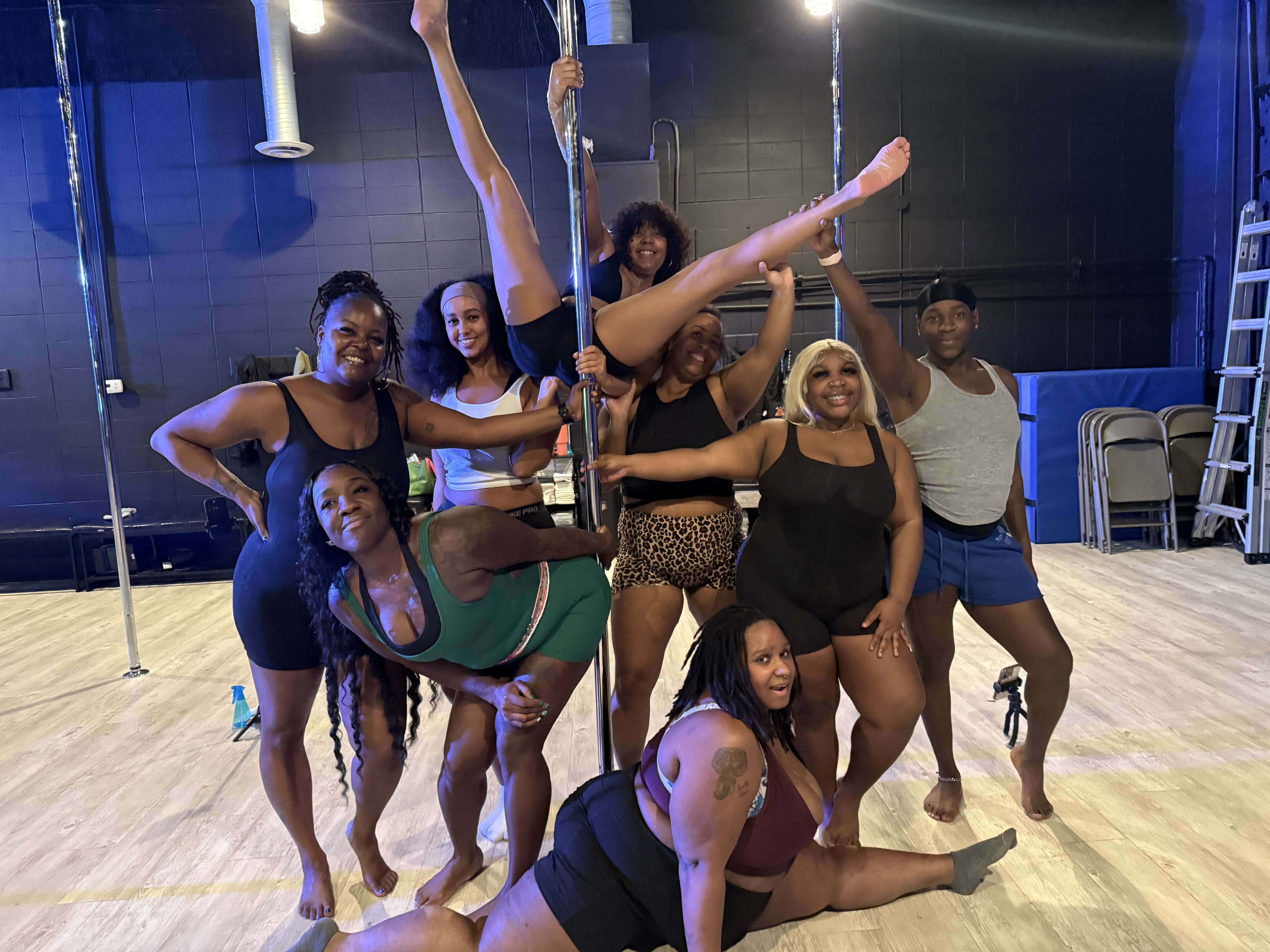 Energetic pole fitness group photo in a dance studio: eight people smiling around a vertical pole, one performing a high split while others strike playful poses on a light wood floor with studio equipment in the background.