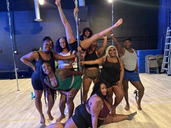 Energetic pole fitness group photo in a dance studio: eight people smiling around a vertical pole, one performing a high split while others strike playful poses on a light wood floor with studio equipment in the background.