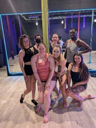 Cheerful group of seven adults posing around a gold dance pole in a neon-lit pole fitness studio — mirrored wall, colorful aerial silks, and string lights in the background.