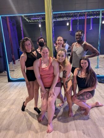 Cheerful group of seven adults posing around a gold dance pole in a neon-lit pole fitness studio — mirrored wall, colorful aerial silks, and string lights in the background.