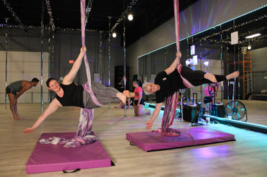 Two adults smiling while suspended horizontally in purple aerial silks over pink crash mats in a mirrored indoor aerial fitness studio with string lights.