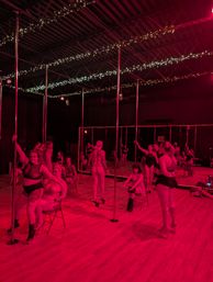 Group pole dance class in a pole fitness studio under red lighting with mirrors, wood floor, vertical poles and string fairy lights