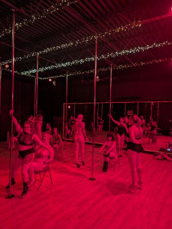 Group pole dance class in a pole fitness studio under red lighting with mirrors, wood floor, vertical poles and string fairy lights