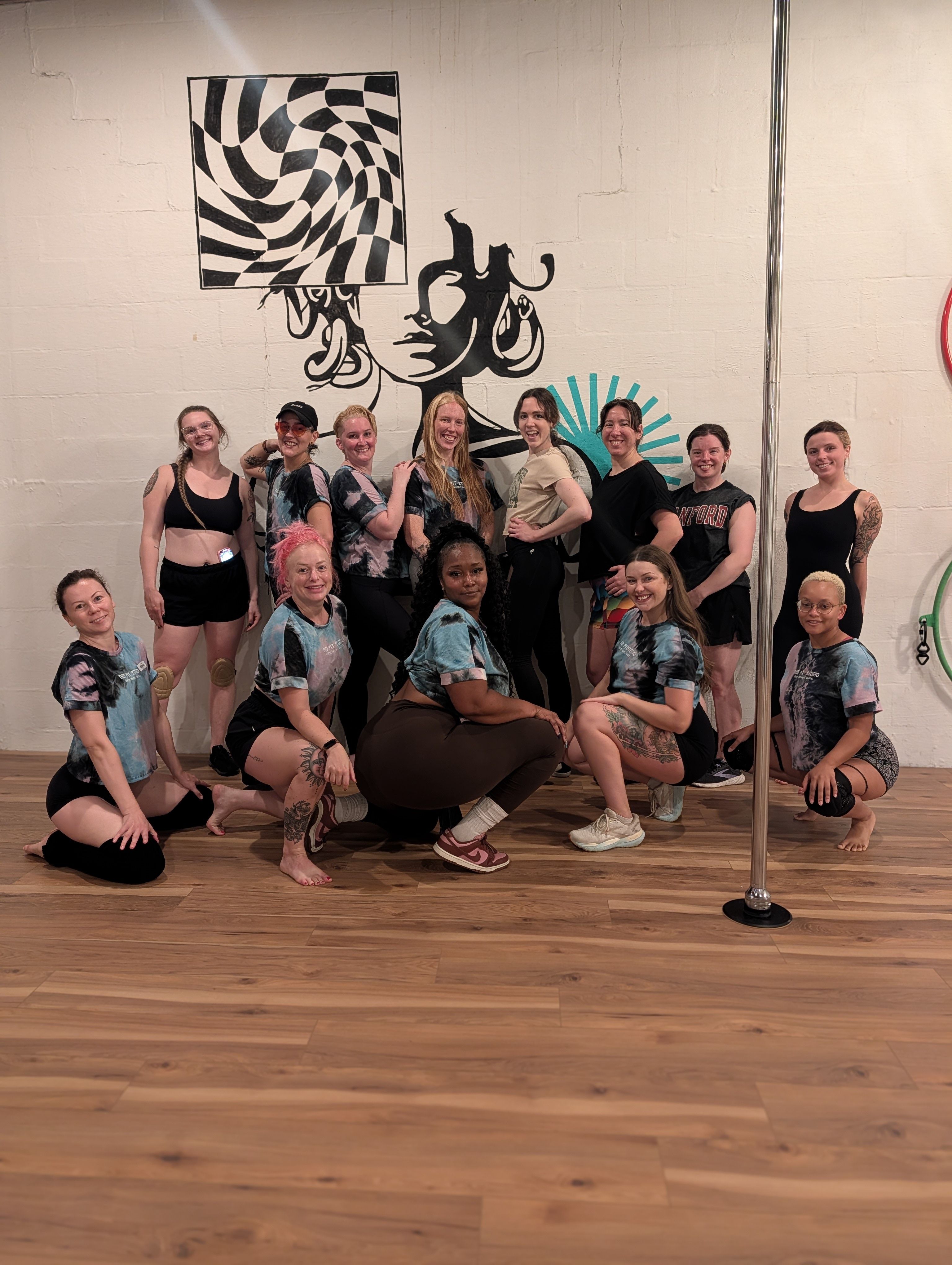 Group photo of a dozen women in athletic wear posing in a pole dance studio on a wood floor, smiling in front of a bold black-and-white mural of a stylized face and geometric pattern with a chrome pole at the right.