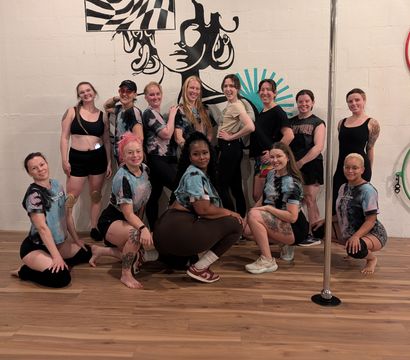 Group photo of a dozen women in athletic wear posing in a pole dance studio on a wood floor, smiling in front of a bold black-and-white mural of a stylized face and geometric pattern with a chrome pole at the right.