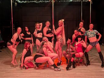 Group of diverse pole dancers posing around a center pole on a studio stage with string lights, wearing pole fitness outfits and high-heeled platform shoes.
