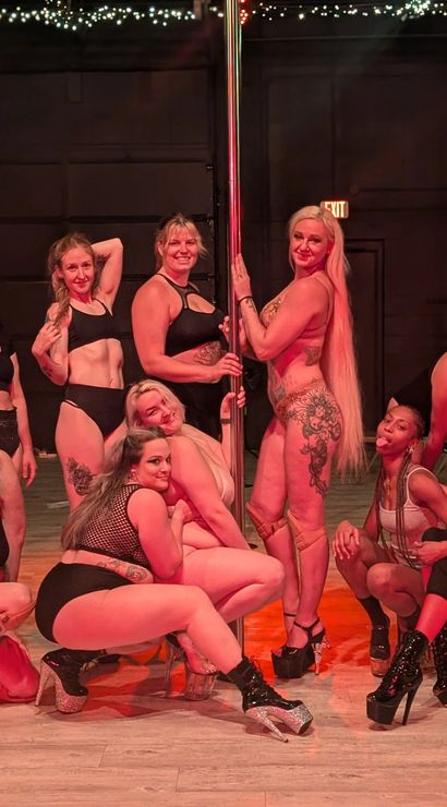 Group of diverse pole dancers posing around a center pole on a studio stage with string lights, wearing pole fitness outfits and high-heeled platform shoes.