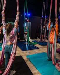 Group aerial silks class in an indoor fitness studio, participants grasp colorful hanging silks above padded mats preparing for aerial yoga and acrobatic practice under studio lights