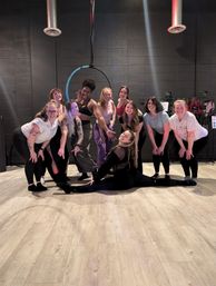 Group of smiling women in athletic wear posing in an indoor aerial hoop fitness studio, one performer doing a full split on the wood floor beneath a suspended hoop against black cinderblock walls.