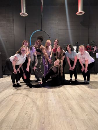 Group of smiling women in athletic wear posing in an indoor aerial hoop fitness studio, one performer doing a full split on the wood floor beneath a suspended hoop against black cinderblock walls.