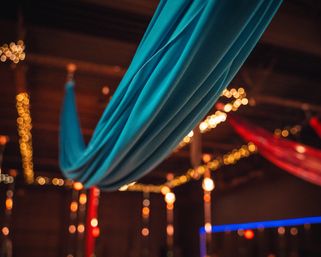 Turquoise ceiling drape in an indoor event venue, soft folds and red fabric accents with warm string lights and glowing bokeh creating cozy ambient lighting.