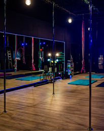 Vibrant pole fitness studio interior with chrome poles, aerial silks and hoops, mirrored wall, teal exercise mats on wood floor, colorful studio lighting and stacked fitness props