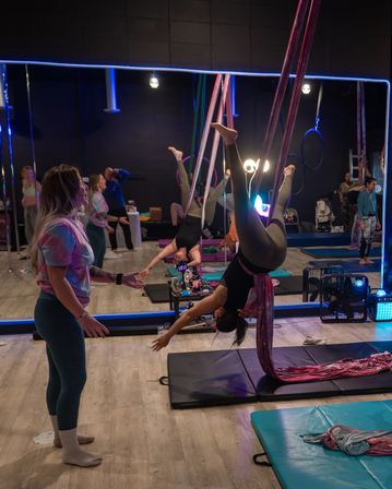 Aerial silks class in an indoor fitness studio — a student suspended upside-down in pink silks over crash mats while an instructor spots, mirrored wall and colorful LED lighting in the background.
