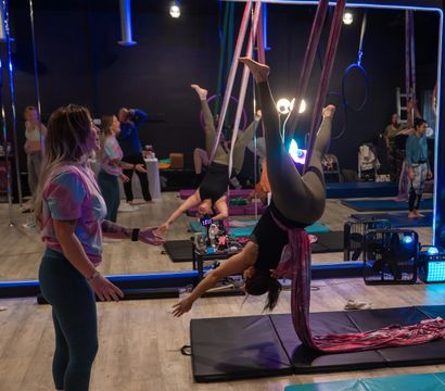 Aerial silks class in an indoor fitness studio — a student suspended upside-down in pink silks over crash mats while an instructor spots, mirrored wall and colorful LED lighting in the background.