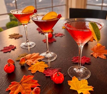 Three red martini cocktails garnished with peach slices and a lime on a dark wooden table scattered with autumn maple leaves, mini pumpkins and acorns, sunset-lit window in background.