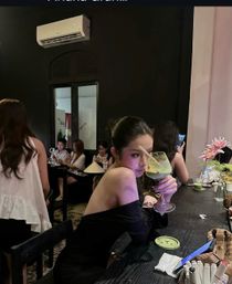 Woman in a black off-shoulder dress sipping a green cocktail from a large glass at a trendy, dimly lit cocktail bar with other patrons socializing in the background.