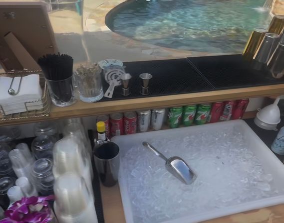 Poolside outdoor bar setup with a large ice bin and metal scoop, cocktail tools and jiggers on a rubber mat, cups, napkins and stirrers, rows of soda cans and metal shakers, with a swimming pool visible in the background.