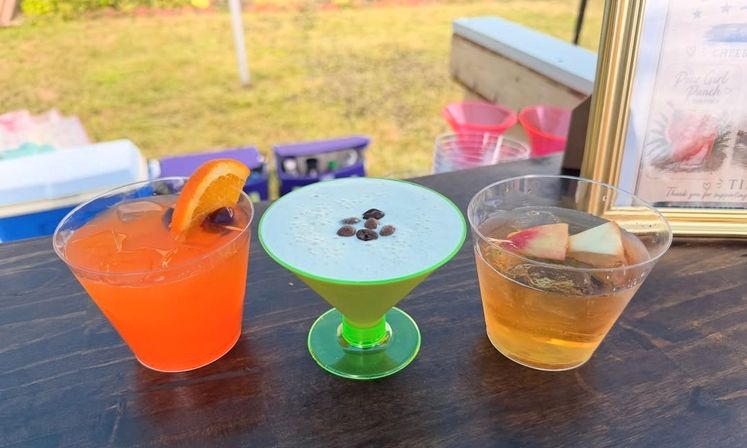Three colorful outdoor summer cocktails on a dark wooden bar: an orange drink with an orange slice and cherry, a frothy blue-green martini topped with coffee beans in a neon green glass, and a golden cocktail garnished with apple slices.