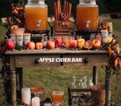 Rustic outdoor apple cider bar in an autumn orchard: wooden table with two large glass cider dispensers, fresh apples lined up, cinnamon sticks, dried citrus slices, mason jars and candles for a cozy fall beverage station.