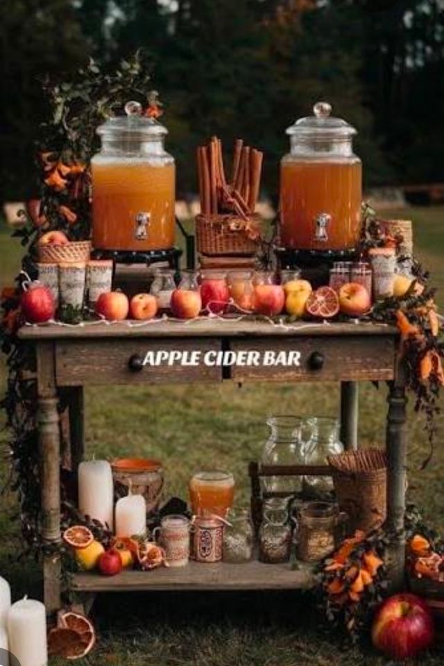 Rustic outdoor apple cider bar in an autumn orchard: wooden table with two large glass cider dispensers, fresh apples lined up, cinnamon sticks, dried citrus slices, mason jars and candles for a cozy fall beverage station.