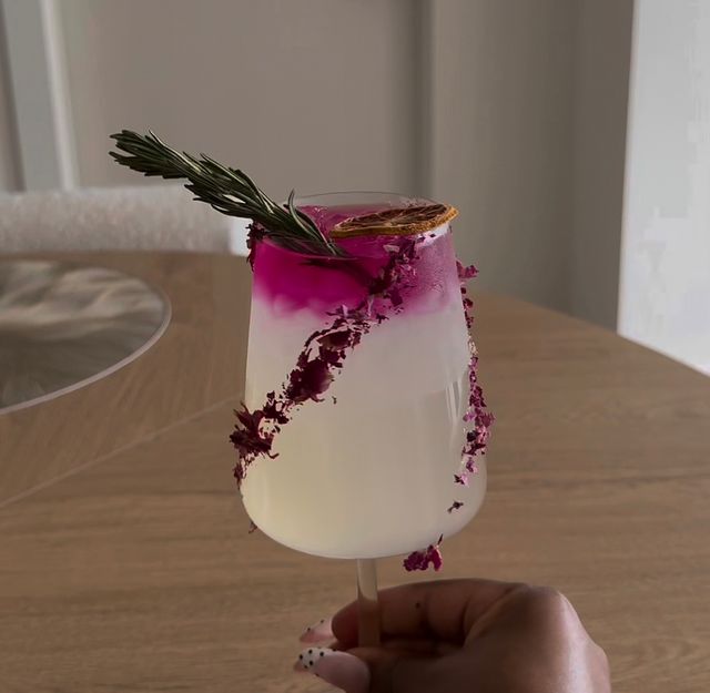 Hand holding an elegant pink-and-white layered cocktail in a stemmed glass garnished with a rosemary sprig, dried citrus slice, and crushed rose petals along the rim on a wooden table