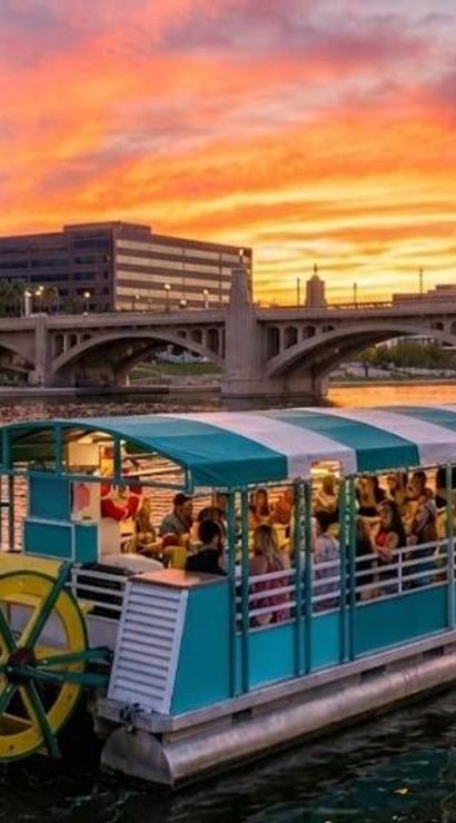 Teal paddlewheel tour boat carrying people on a city river at vibrant orange-pink sunset, with an arched bridge and glass downtown buildings reflected in the water.