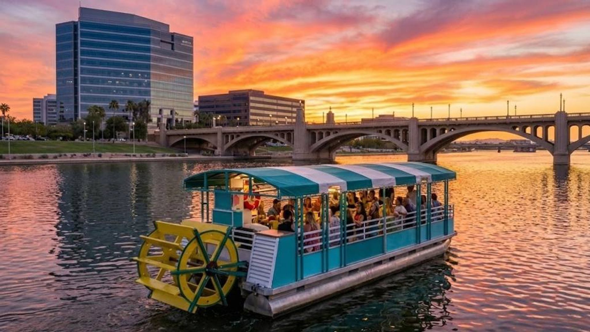Teal paddlewheel tour boat carrying people on a city river at vibrant orange-pink sunset, with an arched bridge and glass downtown buildings reflected in the water.