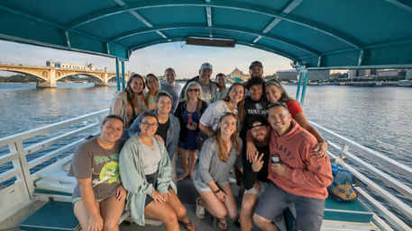 Smiling group of friends on a covered teal sightseeing boat cruising an urban river at golden hour with arched bridge and waterfront skyline