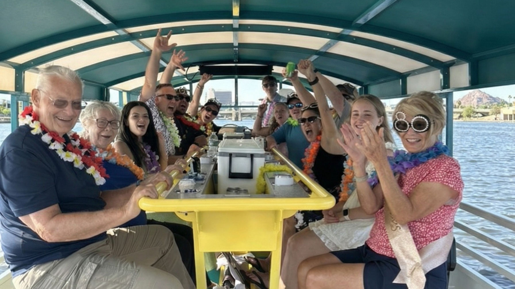 Multigenerational group wearing leis and sunglasses cheering aboard a covered party boat with a yellow bar on a sunny lake cruise