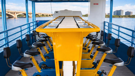 Bright yellow pedal-bar boat with two rows of black bicycle seats on a blue deck, docked on a river with an arched bridge and city skyline in the background — waterfront pedal cruise scene.