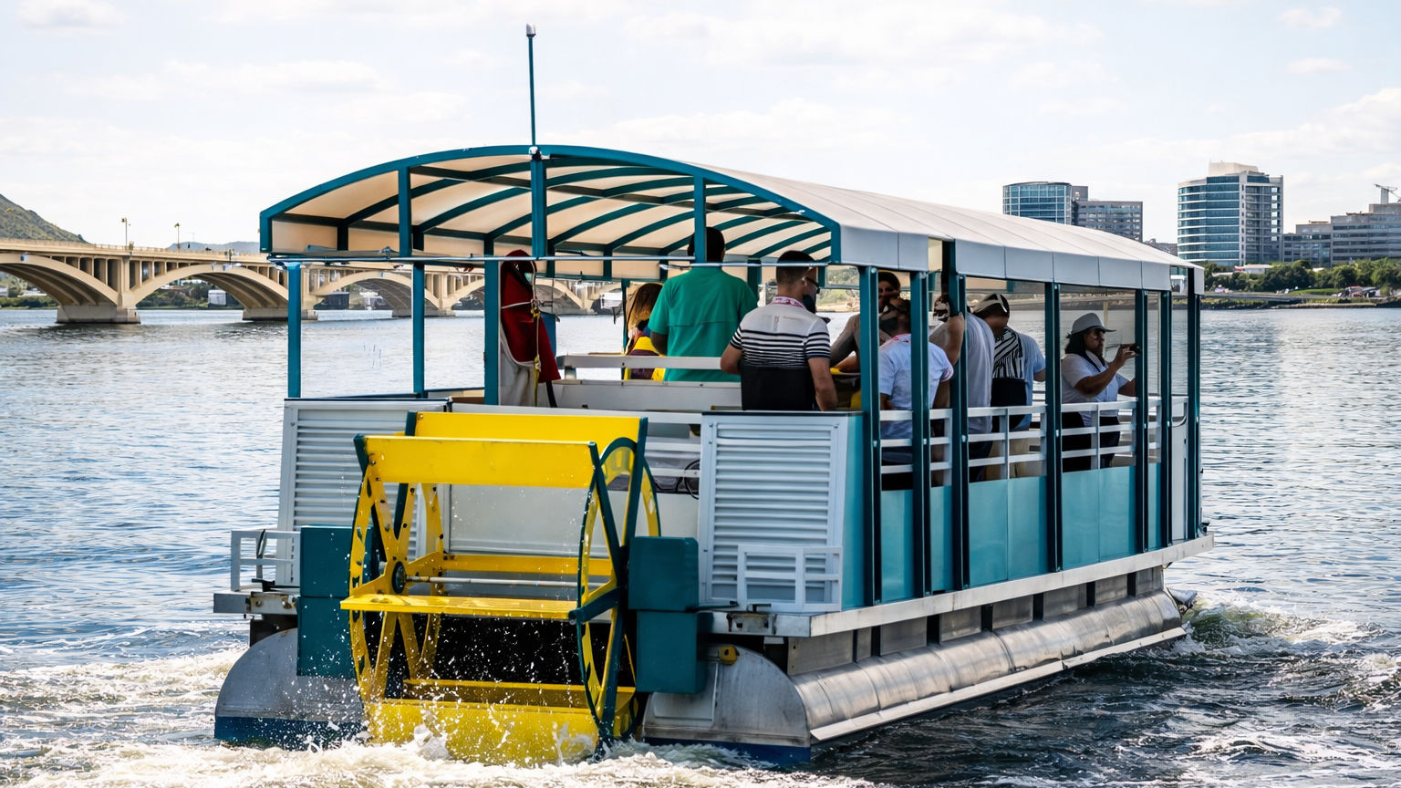 Colorful paddlewheel tour boat with a yellow wheel and covered passenger deck cruising on an urban river, passing an arched bridge and modern city skyline.